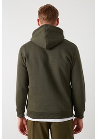 Sudadera verde con una gran capucha, puños acanalados y dobladillo bandado. Textura suave, tejido de peso medio, sobre una camiseta blanca.