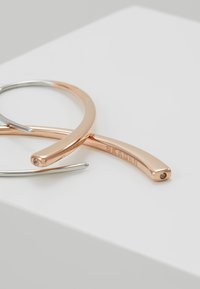 Monture de lunettes en métal au design épuré. Présente un bras argenté et un bras en or rose avec un petit accent en pierre précieuse. Branding gravé présent.