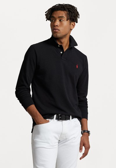 Polo noir à manches longues avec col montant, comportant trois boutons et un petit logo rouge sur la poitrine, associé à un pantalon blanc.