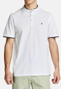 Mand i hvid kortærmet henley-shirt med sort kant på ærmeenderne og tre knapper, parret med lysegrønne bukser.