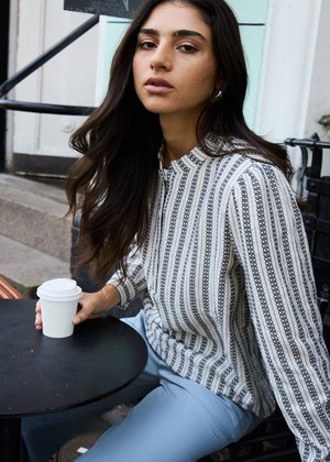 Young woman with long dark hair sitting at outdoor café table, holding a white coffee cup, wearing striped blouse and light blue pants.