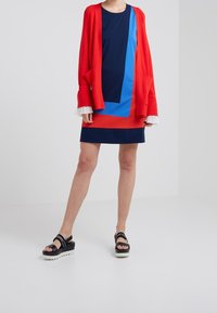Red cardigan over a navy and blue geometric-patterned dress. Black platform sandals with white soles. Textured ribbed sleeve cuffs.