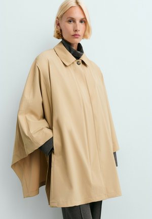 Femme blonde portant un manteau cape beige oversize avec un col roulé noir et un pantalon foncé, debout sur un fond clair.