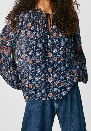 Mujer con una blusa floral azul oscuro holgada de manga larga y vaqueros azules, de pie con las manos en las caderas.