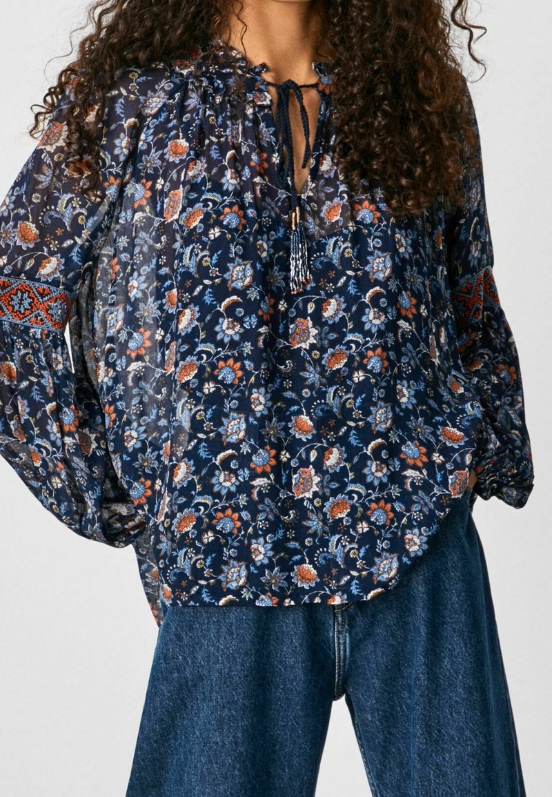 Femme portant un chemisier ample à fleurs bleu foncé avec manches longues et un jean bleu, debout les mains sur les hanches.