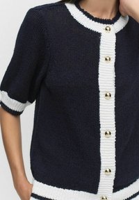 Navyblå stickad cardigan med vit kant, korta ärmar och en strukturerad knappslå med guldfärgade knappar.