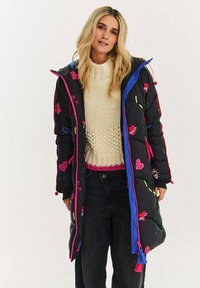 Černý prošívaný kabát s barevným vyšíváním a jasně modrými akcenty. Má přední zip a kapuci, nošený přes krémový pletený svetr a tmavé kalhoty.