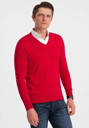 Pull en tricot rouge avec un col en V, des manches longues, des poignets et un ourlet côtelés, arborant un petit logo sur la poitrine. Associé à un jean foncé.