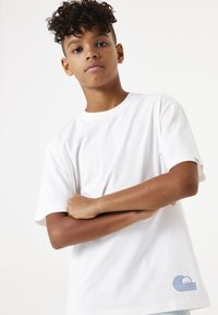 Camiseta de algodón blanca con cuello redondo y mangas cortas, que presenta un diseño de olas azules en la parte inferior izquierda. Textura suave.
