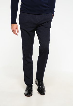 Homme portant un pantalon de costume bleu marine, des chaussures habillées noires et polies, ainsi qu'une chemise manches longues bleu marine, debout avec une main dans la poche.