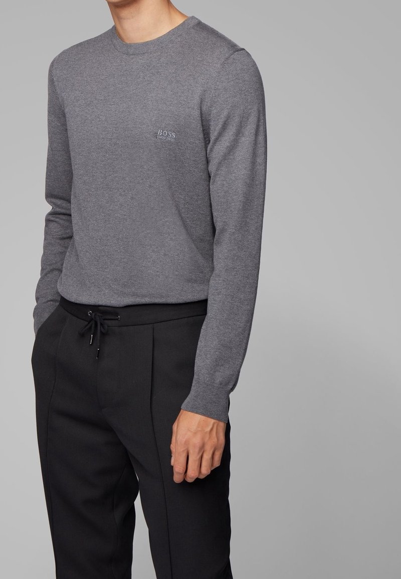 Grauer Langarmpullover mit rundem Ausschnitt und weißem Logo auf der linken Brust, kombiniert mit schwarzen Jogginghosen mit Kordelzug. Glatte Textur.