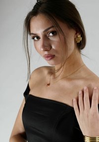 Svart off-shoulder klänning, gyllene spiralörhängen, delikat gyllene halsband med en röd ädelsten, slät hud, naturlackade naglar, neutral bakgrund.