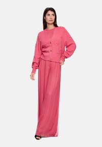 Rosa gestrickte Strickjacke mit floralen Verzierungen und goldenen Knöpfen, getragen über einem passenden langen plissierten Kleid, ergänzt durch schwarze Absätze.