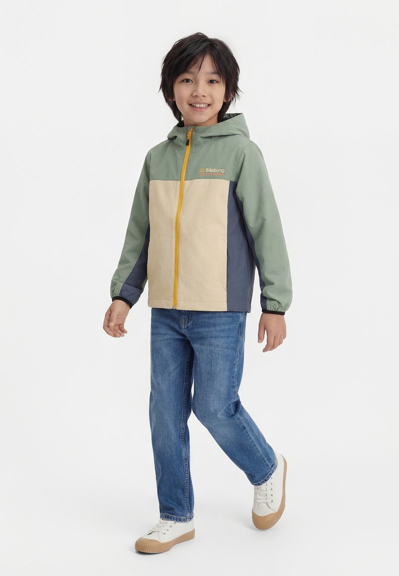 Dieťa v zeleno-béžovo-modrej bunde s kapucňou, modrých džínsoch a bielych teniskách kráča a usmieva sa na bielom pozadí.