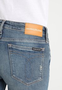 Jeans de mezclilla azul con una etiqueta de cuero color tan en la pretina que muestra "CALVIN KLEIN JEANS." Costuras visibles, bolsillos traseros y un ligero desgaste.