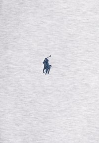 La tela de algodón gris presenta un pequeño logotipo bordado de un jugador de polo en azul oscuro en la parte izquierda del pecho. La textura parece suave y ligeramente jaspeada.