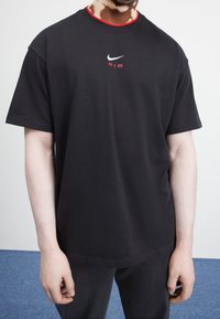 Person wearing black Nike Air t-shirt and black pants, standing against a white and blue background.
