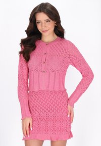 Femme portant un haut à manches longues en tricot rose avec boutons et une jupe assortie aux motifs complexes en dentelle, posant avec une main sur la hanche.