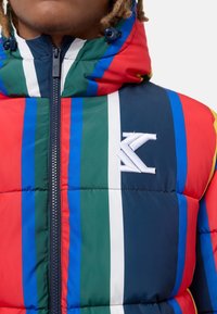 Giacca imbottita con strisce audaci in rosso, verde, blu e navy, con chiusura a zip e logo ricamato sul petto.