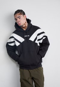 Svart sweatshirt med vita diagonala ränder och logotyp. Blandat bomullsmaterial, normal passform, känguruficka, justerbar dragsko på huvan.