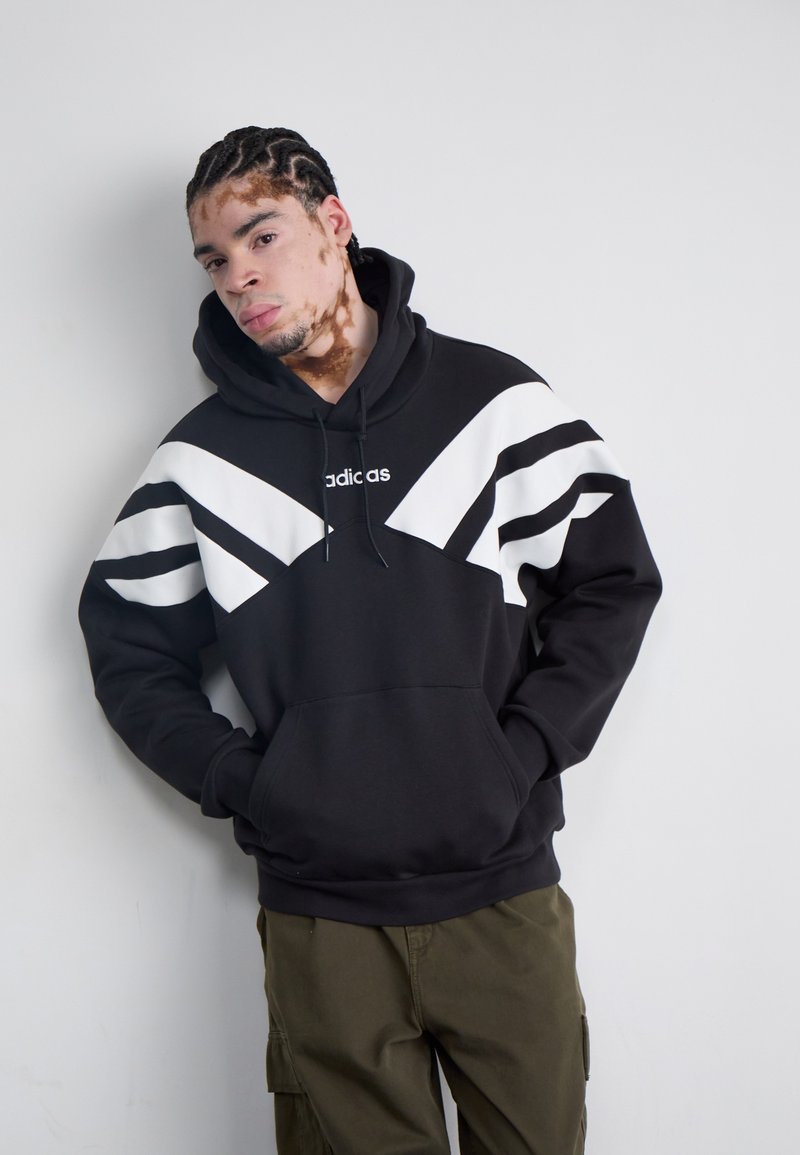 Svart sweatshirt med vita diagonala ränder och logotyp. Blandat bomullsmaterial, normal passform, känguruficka, justerbar dragsko på huvan.
