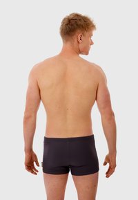 Shorts de bain noirs avec une texture lisse et une coupe ajustée. Le modèle est montré de dos avec des cheveux courts et une silhouette athlétique.