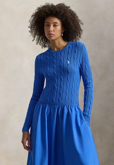Μπλε πουλόβερ με πλέξη καλώδιο, μακριά μανίκια και στρογγυλή λαιμόκοψη. Το τμήμα της φούστας σε μονόχρωμο ύφασμα μπλε, δημιουργώντας ένα στρωματοποιημένο αποτέλεσμα.