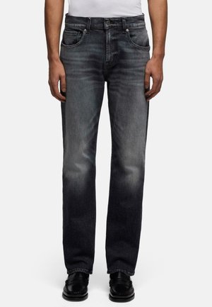 Mand iført falmede sorte jeans med lige ben og fem lommer samt sorte lædersko, stående mod en hvid baggrund.