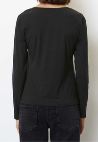 Kvinna iklädd en enkel svart långärmad topp och mörka jeans, visad bakifrån mot en neutral bakgrund.