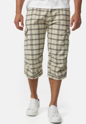 Shorts cargo à carreaux beiges avec des rayures noires, long au genou, dotés de poches latérales. Associés à des baskets blanches pour un look décontracté.