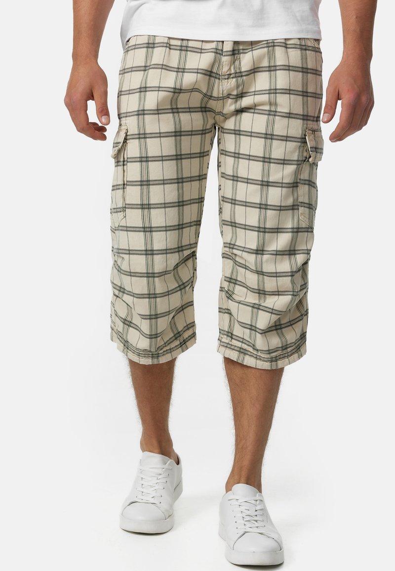 Shorts cargo à carreaux beiges avec des rayures noires, long au genou, dotés de poches latérales. Associés à des baskets blanches pour un look décontracté.