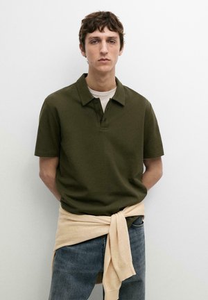 Jeune homme portant un polo vert olive avec un pull beige noué autour de la taille et un jean bleu, debout devant un fond blanc.