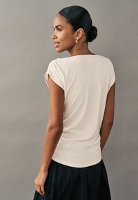 Mujer con cabello oscuro en moño, vestida con top blanco de manga corta y falda negra, de perfil con fondo gris neutro.