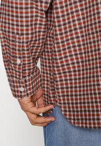 Camisa de franela a cuadros en marrón, rojo y blanco. Presenta un dobladillo curvado, puños abotonados y una textura suave con detalles de costura visibles.