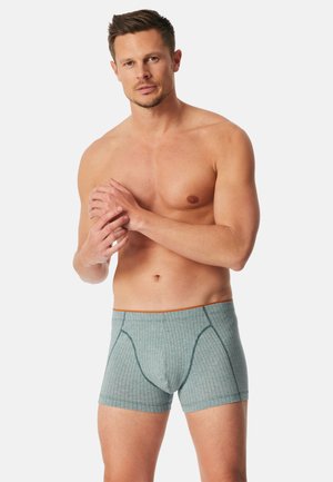 Oberkörperfreier Mann mit kurzen Haaren, der graue gestreifte Boxershorts trägt und vor einem einfarbigen hellen Hintergrund mit verschränkten Händen steht.
