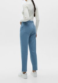 Pantalon en velours côtelé bleu clair, taille haute, jambes droites, avec deux poches à l'arrière, porté avec des baskets blanches sur un fond uni.