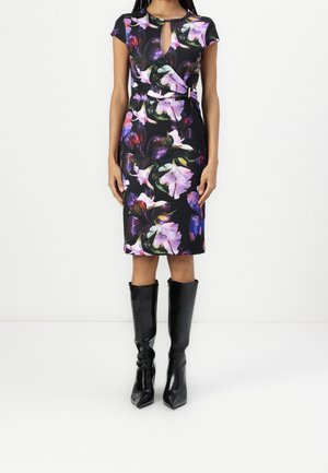 Femme portant une robe noire fleurie mi-longue avec une ouverture en forme de trou de serrure à l'avant et un détail portefeuille, associée à des bottes noires à talons montant jusqu'au genou.