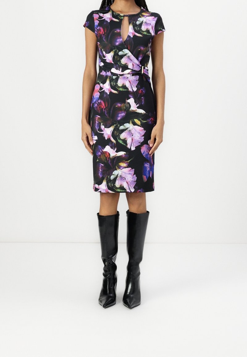 Femme portant une robe noire fleurie mi-longue avec une ouverture en forme de trou de serrure à l'avant et un détail portefeuille, associée à des bottes noires à talons montant jusqu'au genou.