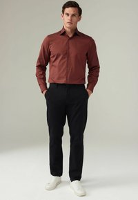 Chemise rouge à manches longues, pantalon noir et baskets blanches portés par une silhouette masculine debout sur un fond neutre.