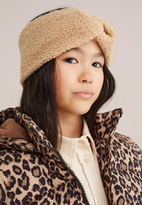 Beige flīsa galvassega ar savērptu mezglu, ko valkā bērns ar gariem tumšiem matiem, kopā ar leoparda raksta jaki.