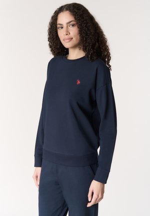 Donna con capelli ricci che indossa una felpa blu navy e pantaloni abbinati, in piedi contro uno sfondo chiaro e uniforme.