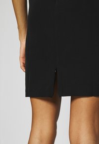 Femme portant une jupe noire avec une fente centrale au dos, montrant la partie inférieure de la cuisse et une partie du bras gauche sur un fond uni.