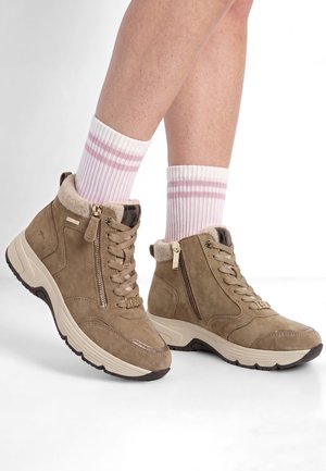 Jambes portant des baskets montantes beiges avec des fermetures éclair latérales et des chaussettes blanches avec deux rayures roses, debout sur un sol blanc.