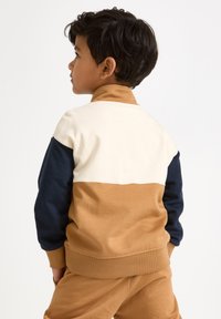 Sötét hajú fiú balra néz, színblokkolt pulóvert visel, amely krémszínű, tengerészkék és bézs színű részekből áll, valamint bézs nadrágot a fehér háttér előtt.