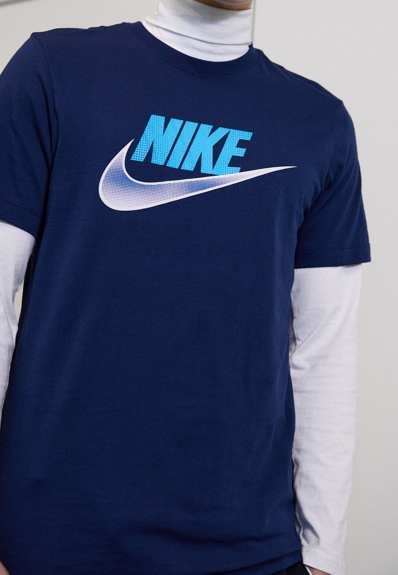 Mörkblå bomulls T-shirt med en stor, texturerad ljusblå "NIKE"-logotyp och en vit swoosh-design, bärs över en vit långärmad skjorta.