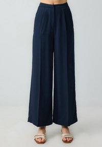 Marineblauwe wijde broek met hoge taille, gemaakt van een gladde stof. Voorzien van plooien aan de voorkant en zijzakken, gecombineerd met neutrale slide sandalen.