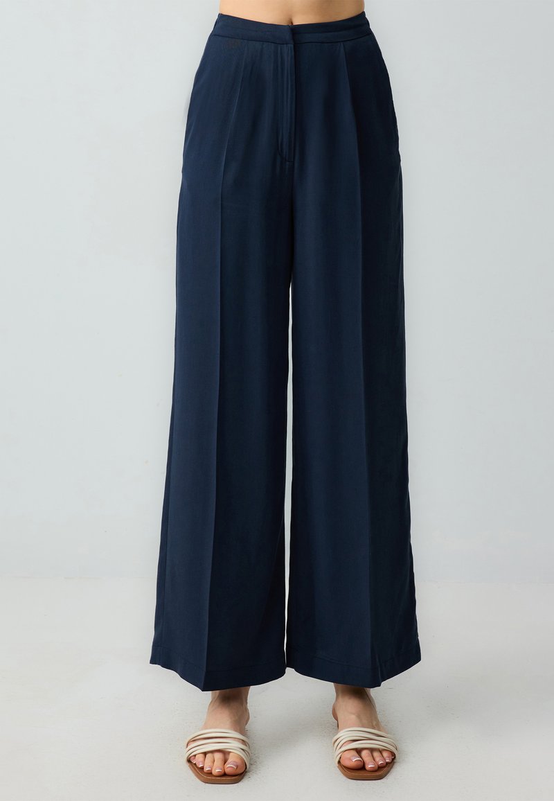 Marineblauwe wijde broek met hoge taille, gemaakt van een gladde stof. Voorzien van plooien aan de voorkant en zijzakken, gecombineerd met neutrale slide sandalen.