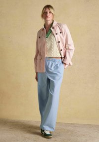 Kvinde iført lyserød jakke, cremefarvet kabelstrikket sweater med grøn kant, blå, bredbenede bukser med striber og grønne sneakers, stående ved en beige væg.