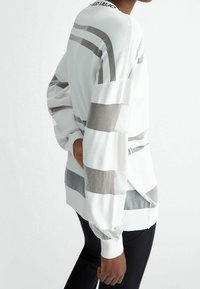 Maglione bianco lavorato a maglia caratterizzato da strisce verticali grigie, pannelli trasparenti, polsini a costine e un logo sul collo sul retro. Tessuto morbido, vestibilità comoda.