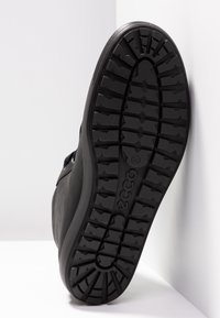 Semelle noire de chaussure ECCO avec des crampons profonds, inclinée contre une surface blanche, montrant le motif d'adhérence et le logo de la marque.